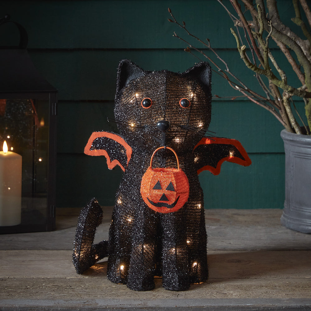Leuchtende Halloween-Deko Fledermaus-Katze mit orangefarbenem Kürbiskörbchen vor dunklem Hintergrund