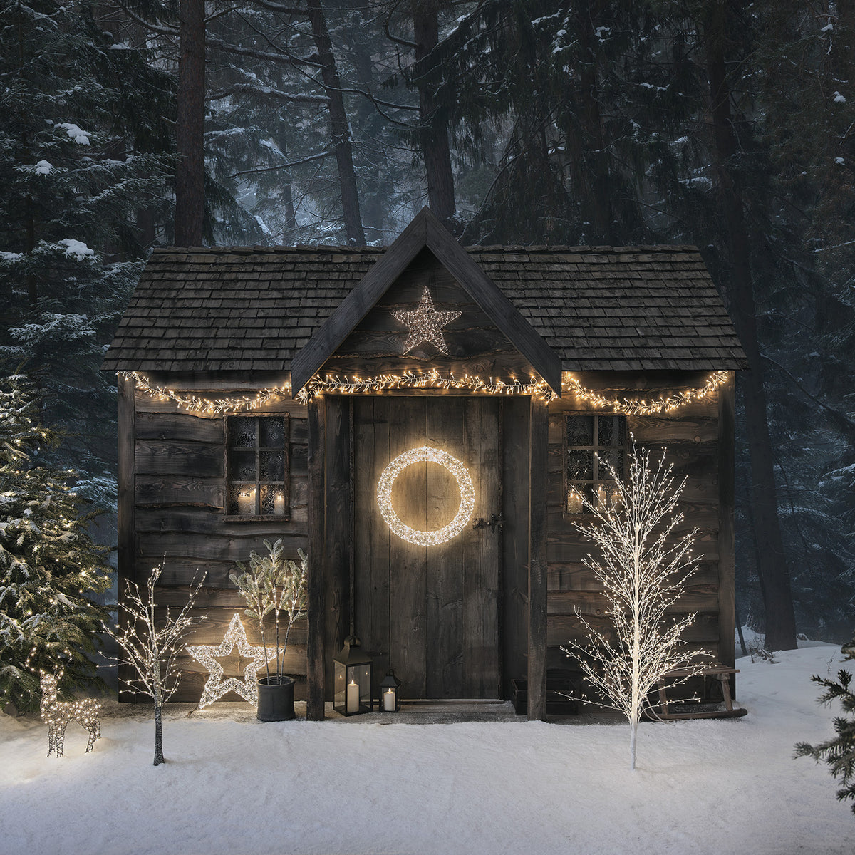Winterliche Hütte mit Weihnachtsbeleuchtung, beleuchteter Stern, Kranz und zwei weißen LED-Draht-Bäumen im Schnee