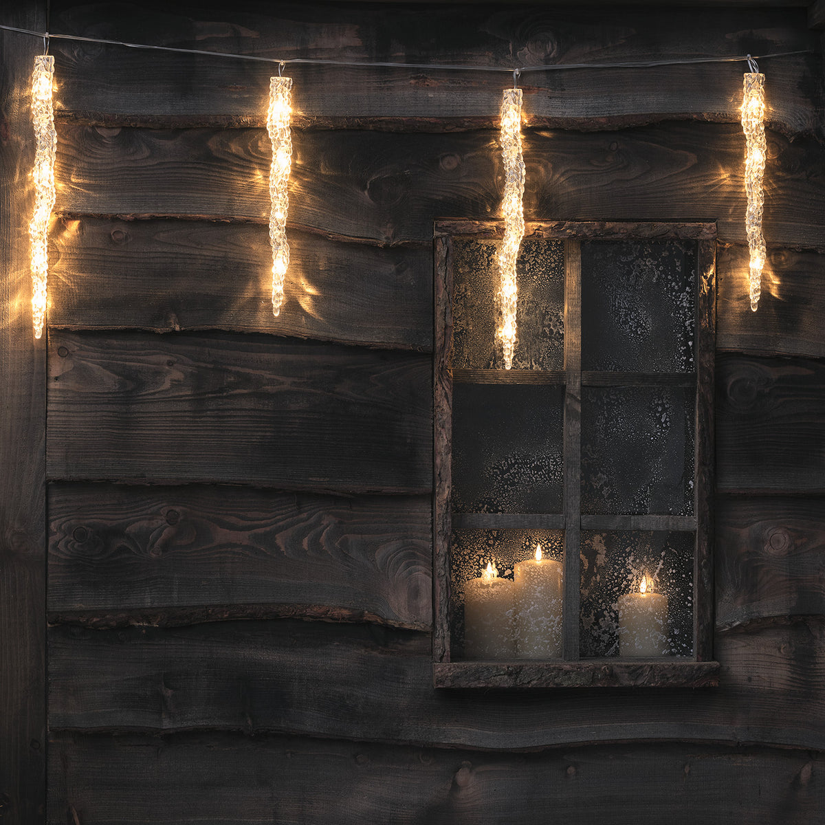 Warmweiße LED Eiszapfen-Lichterkette an dunkler Holzfassade mit Kerzen hinter Fenster