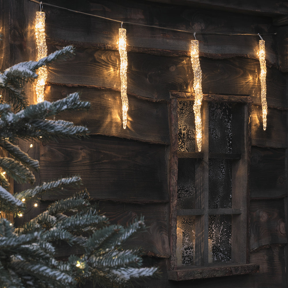 LED Eiszapfen-Lichterkette an Holzhaus mit verschneitem Tannenbaum im Vordergrund