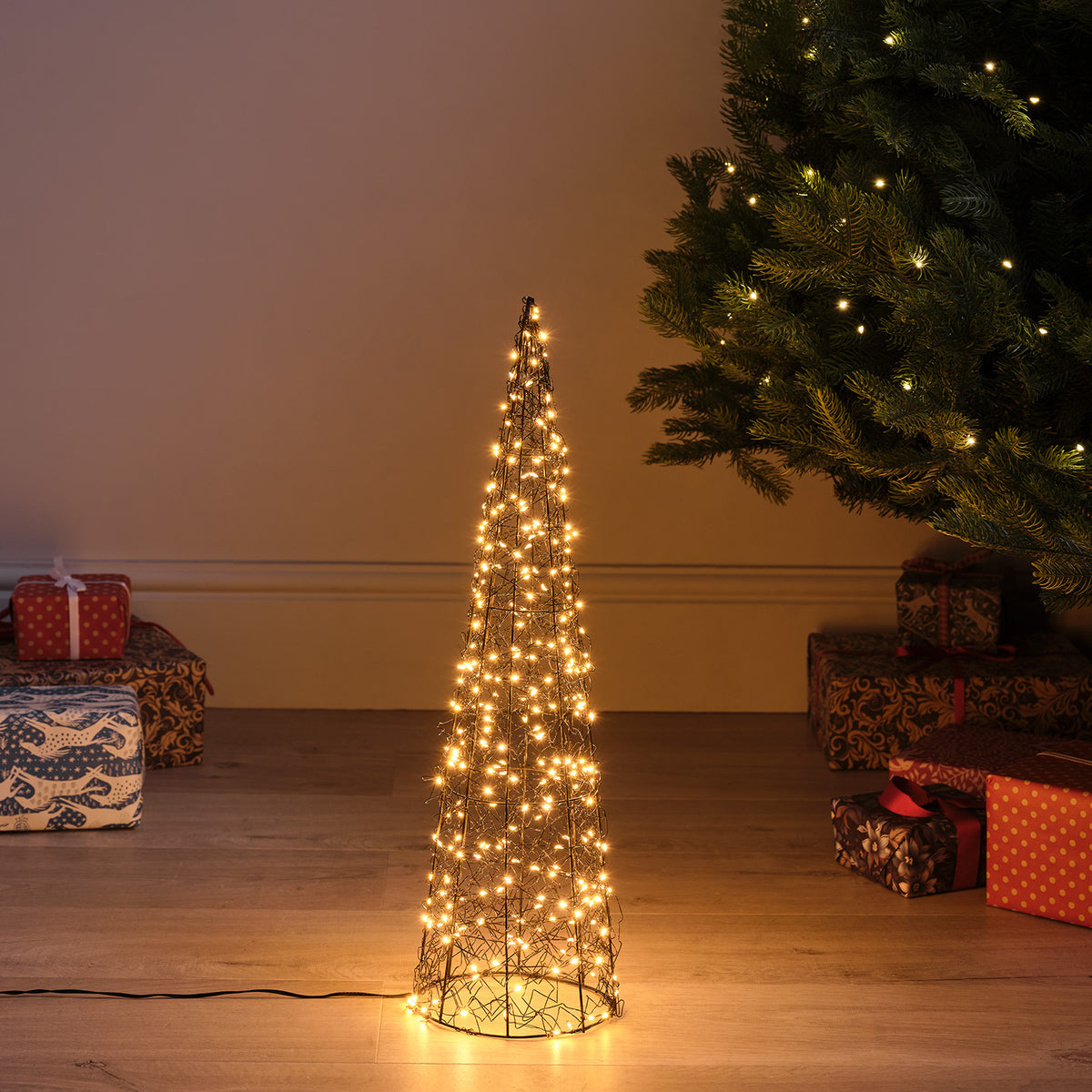Beleuchteter LED-Kegel aus Metalldraht mit warmweißen Micro-LEDs vor Wand und Weihnachtsbaum