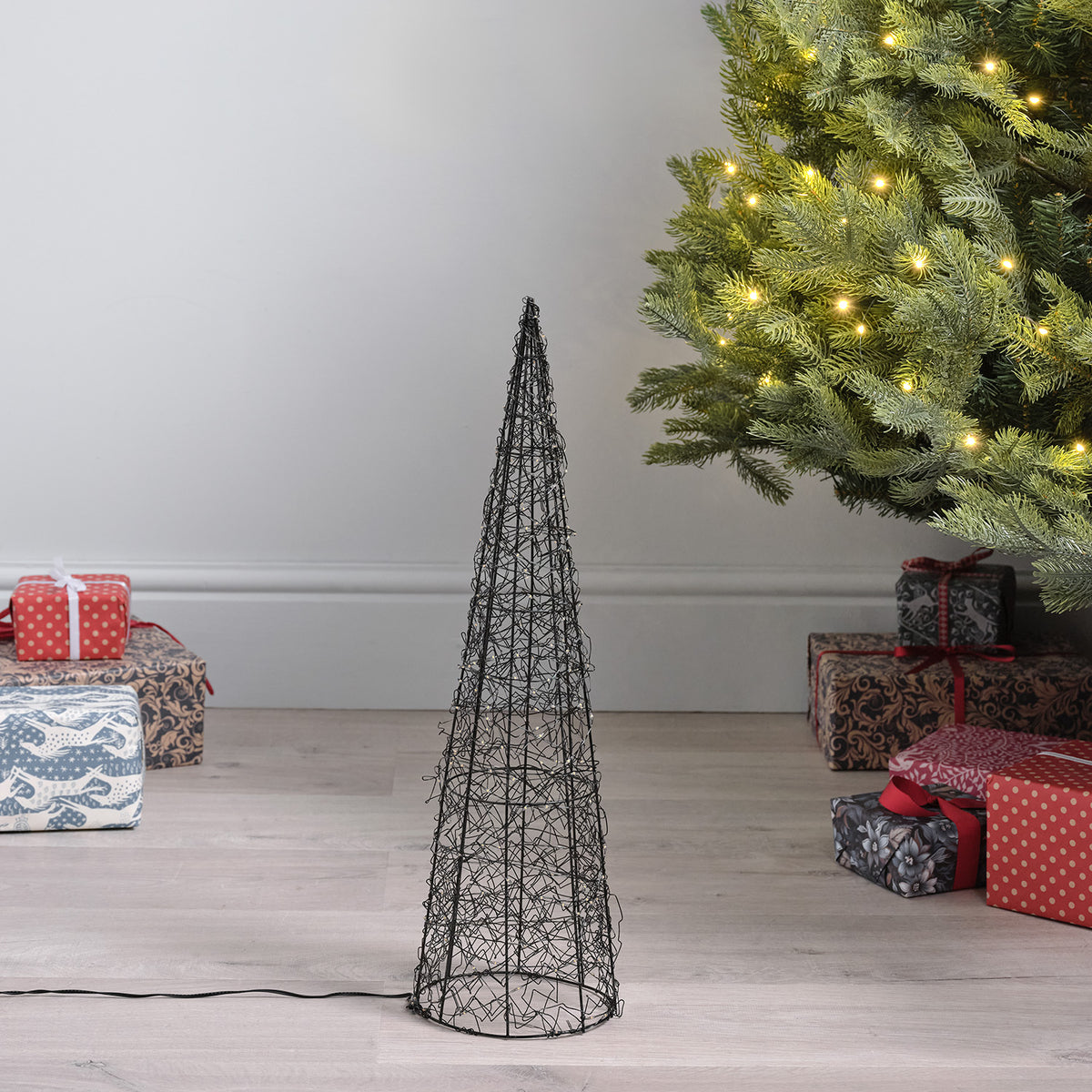 Unbeleuchtete LED-Kegel aus Metalldraht in zwei Größen vor Weihnachtsbaum und Geschenken auf Holzboden