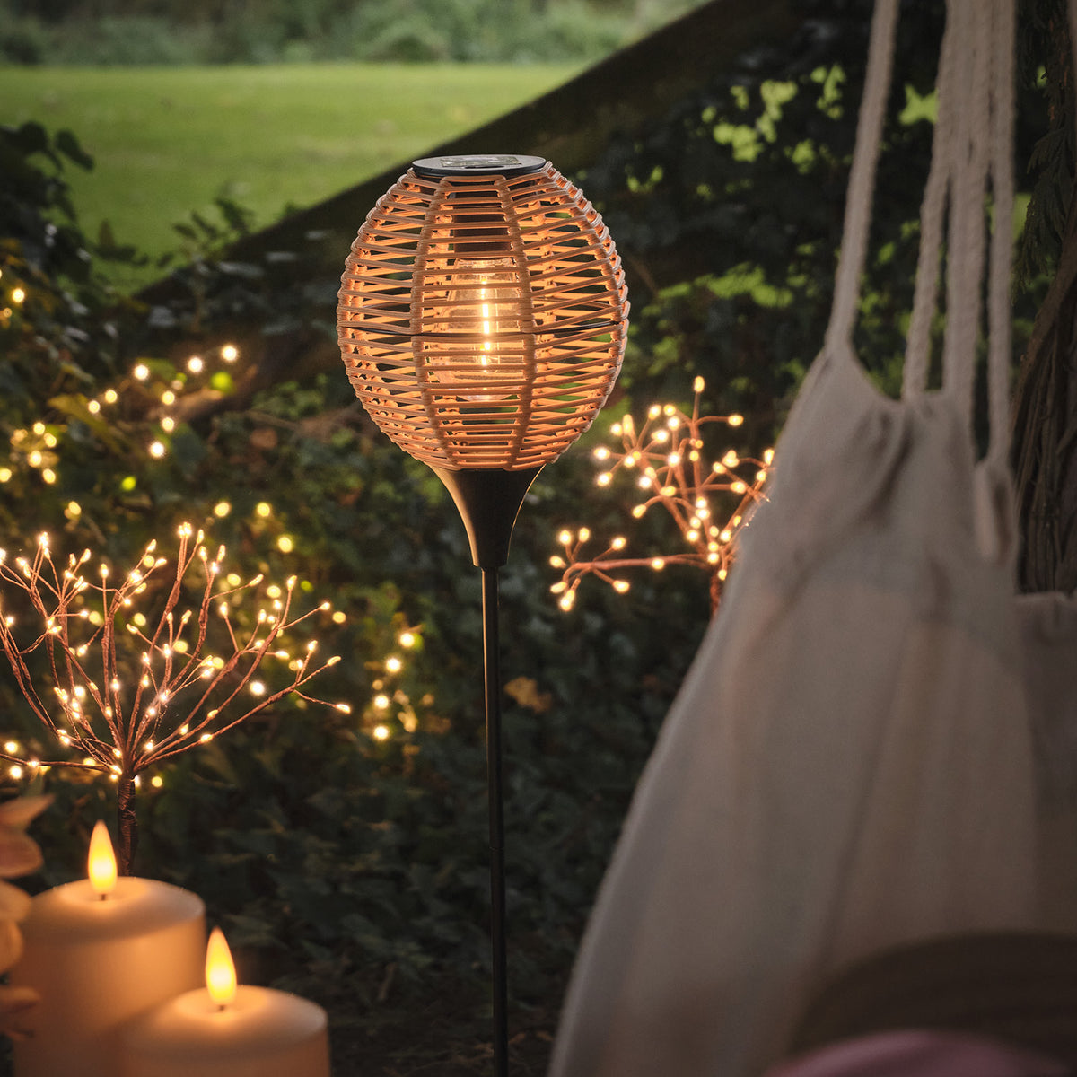 Solar-Gartenstecker in Rattan-Optik leuchtet warmweiß neben Pflanzen und Kerzen im Garten