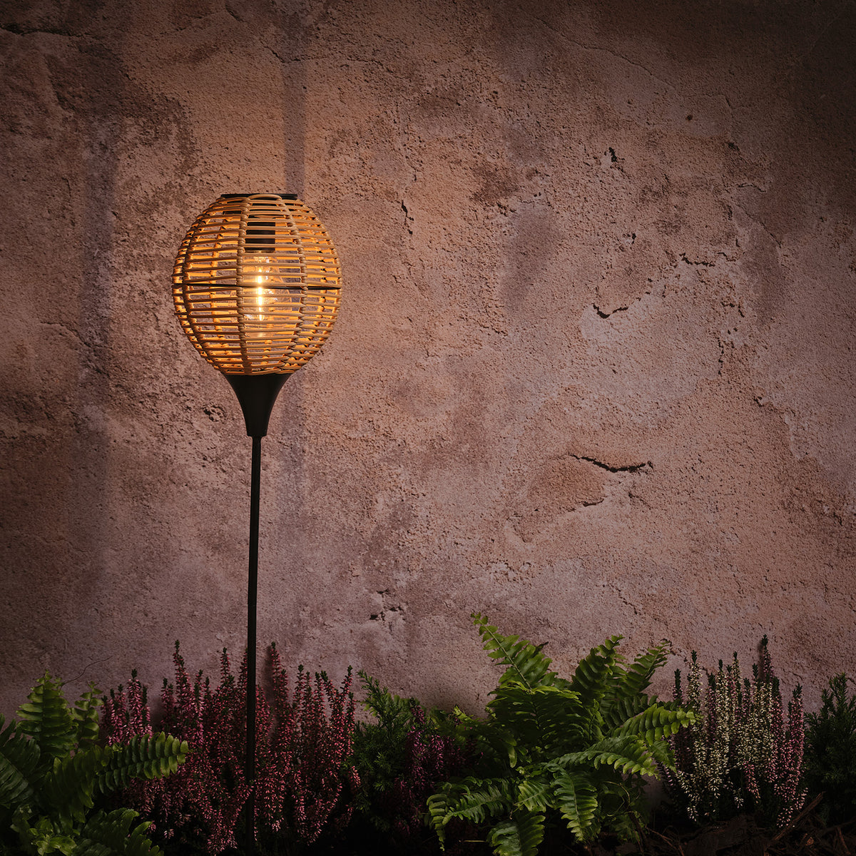 Solar-Gartenstecker mit warmweißem Licht und Rattan-Schirm vor Wand und Heidepflanzen