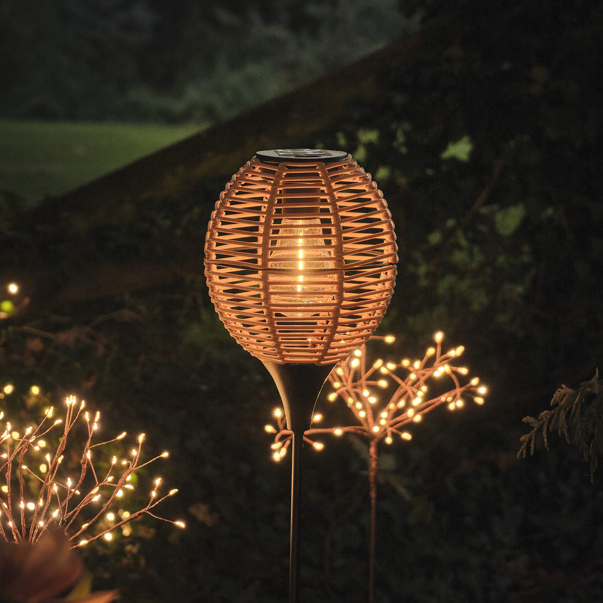 Solar-Stableuchte mit warmweißem Licht in Flechtoptik, umgeben von weiteren Lichterketten im Garten