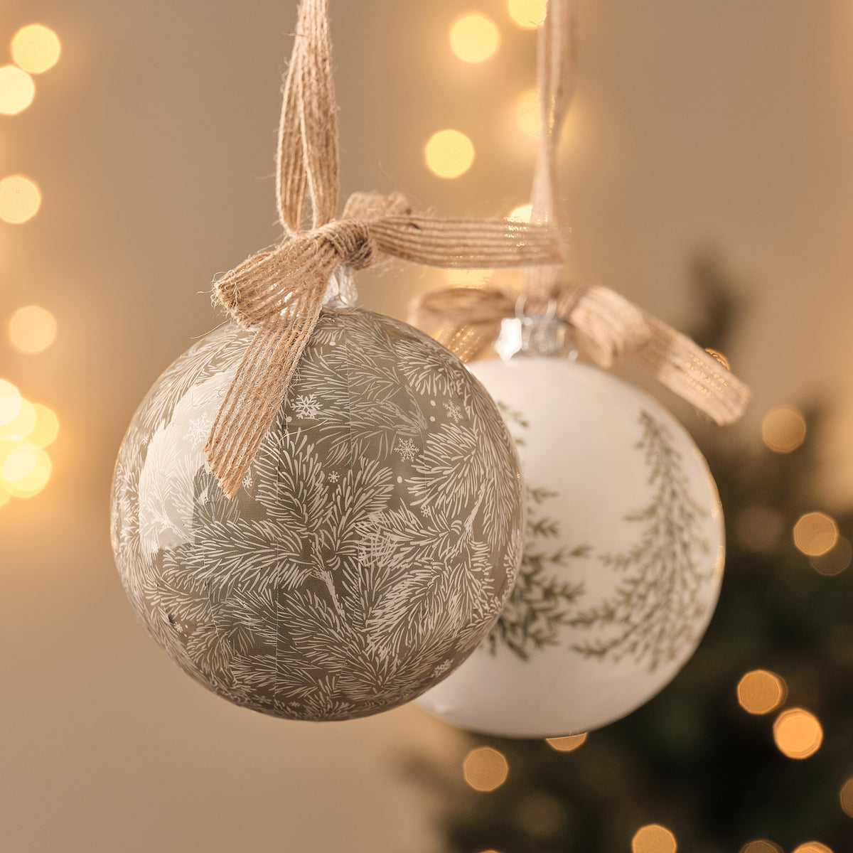 Weiße und goldene Weihnachtskugeln mit Tannenzweig-Motiv und Jute-Schleife, hängend vor Lichterglanz.