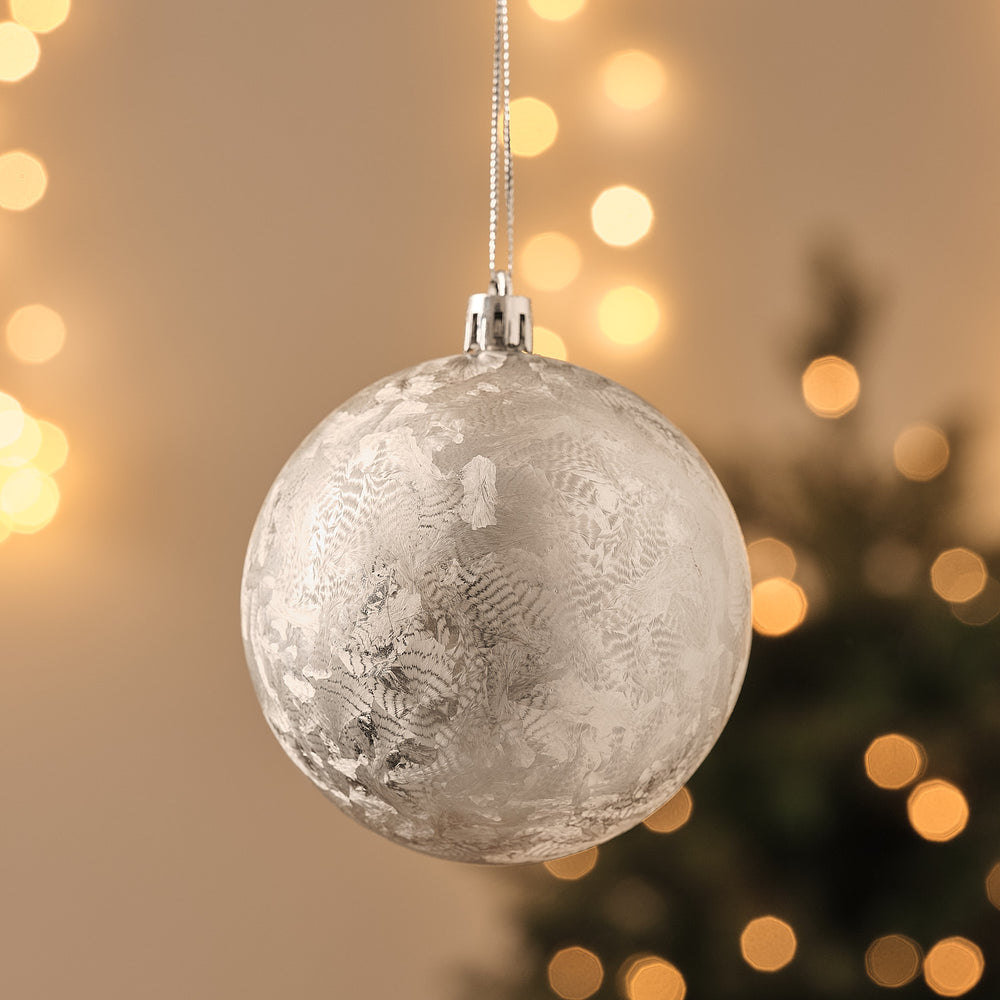 Silberne Weihnachtskugel mit eisblumenartigem Schneeflockenmuster, hängend vor Lichterglanz.