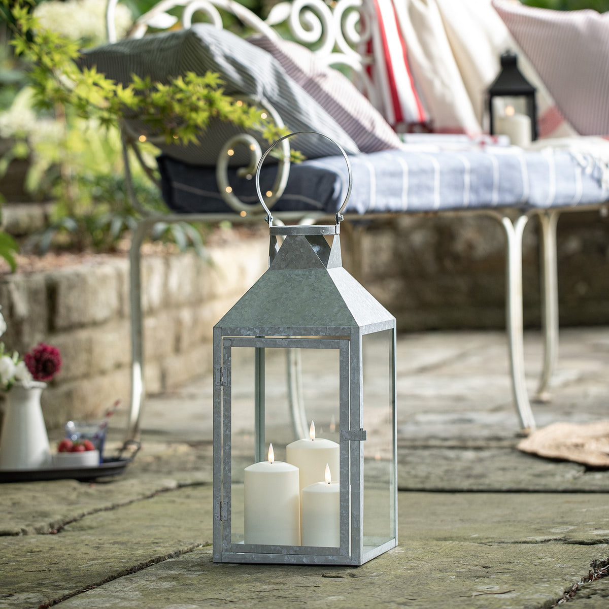 Große verzinkte Outdoor-Laterne mit drei TruGlow LED-Kerzen auf Steinboden vor Gartenbank