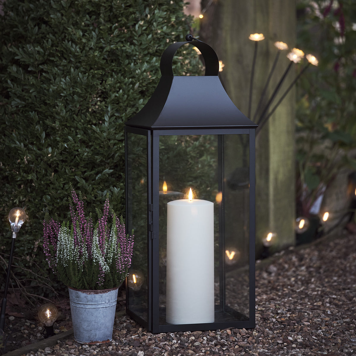 Gartenlaterne mit TruGlow Outdoor Kerze neben Blumentopf mit Heidepflanzen im Außenbereich