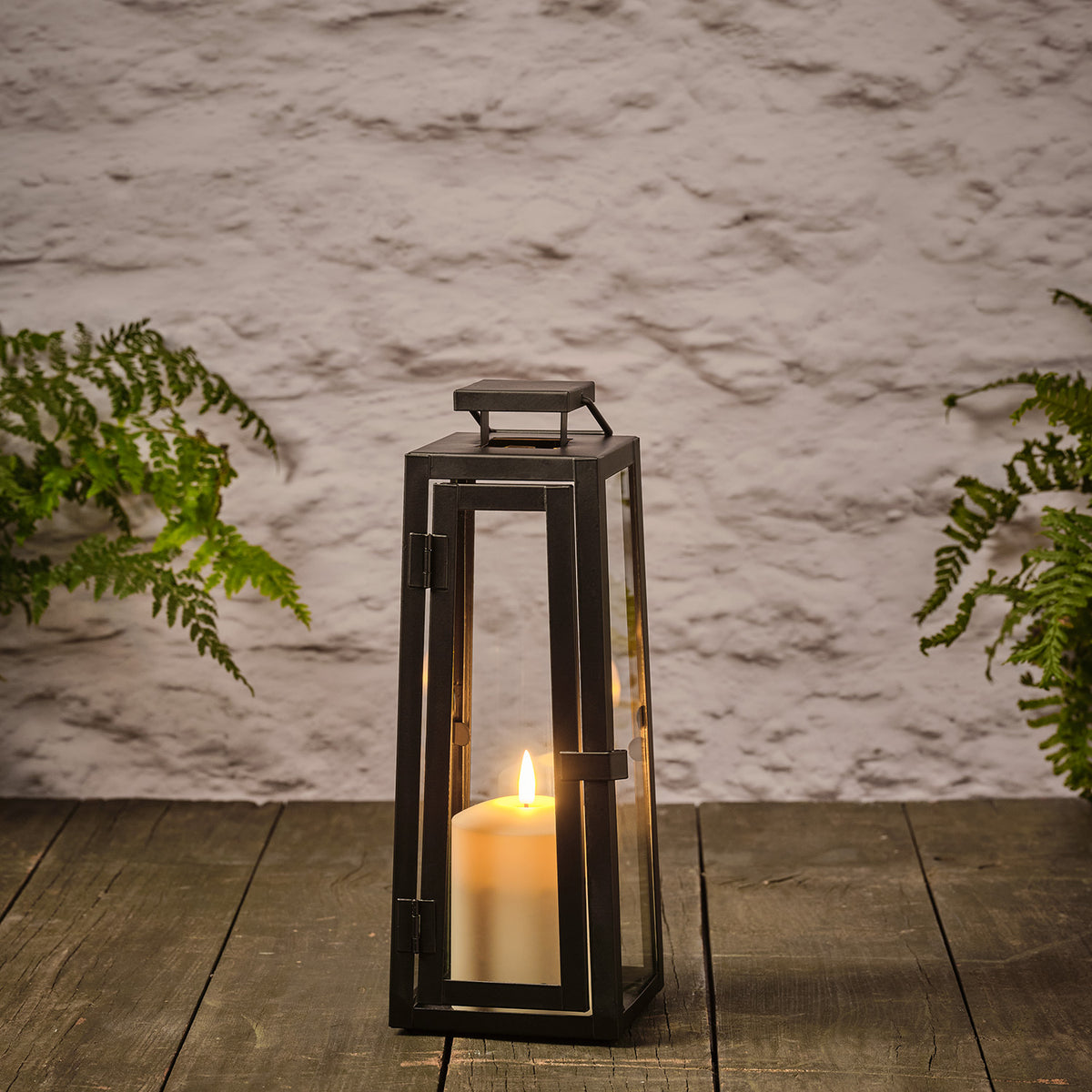 Schwarze Metalllaterne mit Glas und TruGlow LED-Kerze auf Holz vor weißer Wand mit grünen Farnen