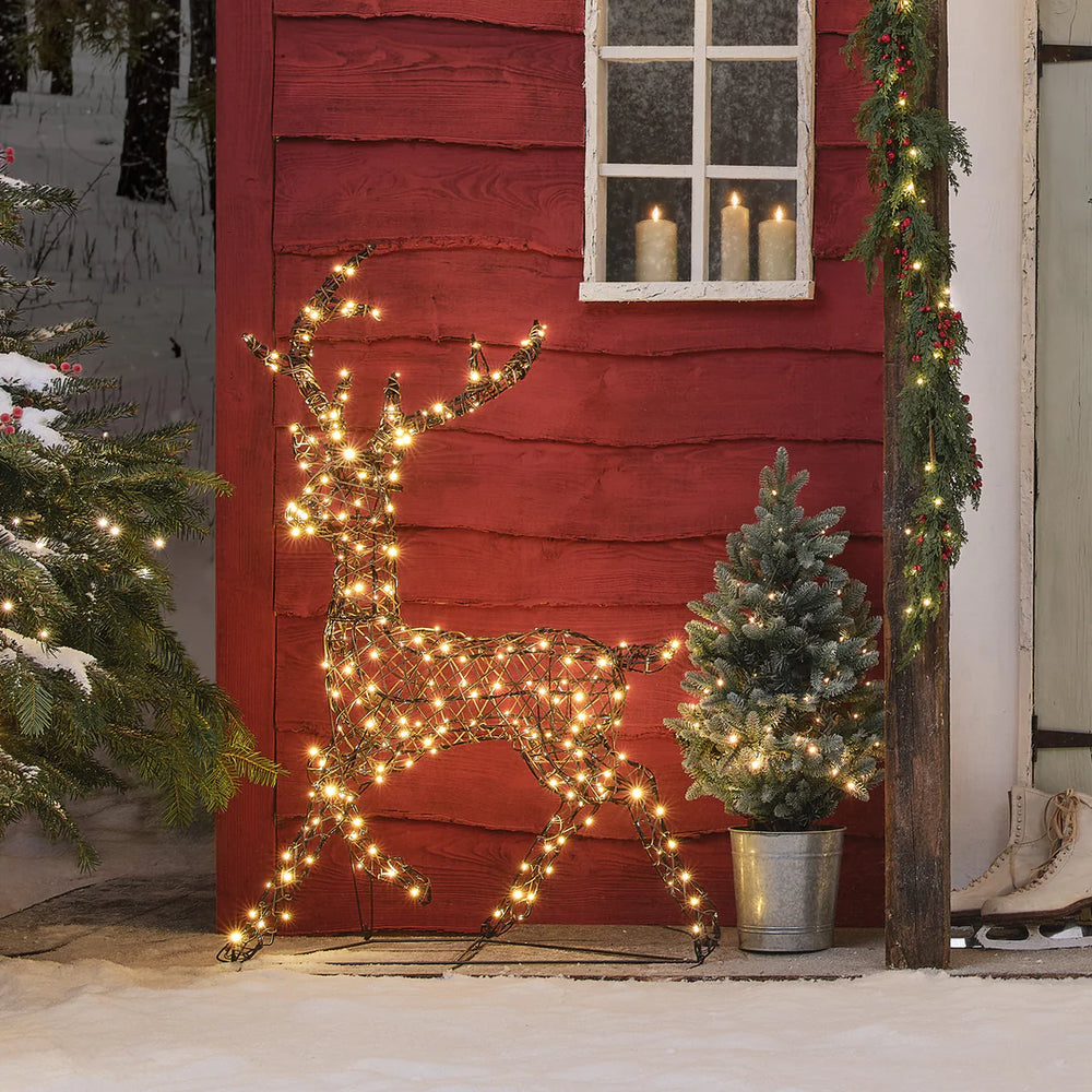 Leuchtendes LED-Rentier in warmweiß neben Tannenbaum und rotem Haus