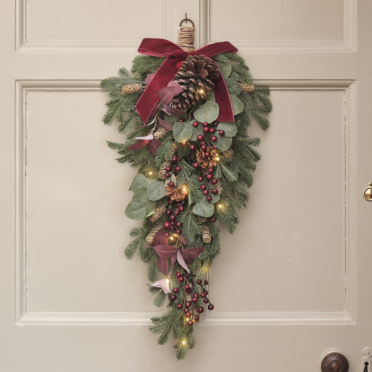 60 cm langer LED-Weihnachtszweig mit roten Beeren, Zapfen, grünen Blättern und Samtschleife an einer hellen Tür