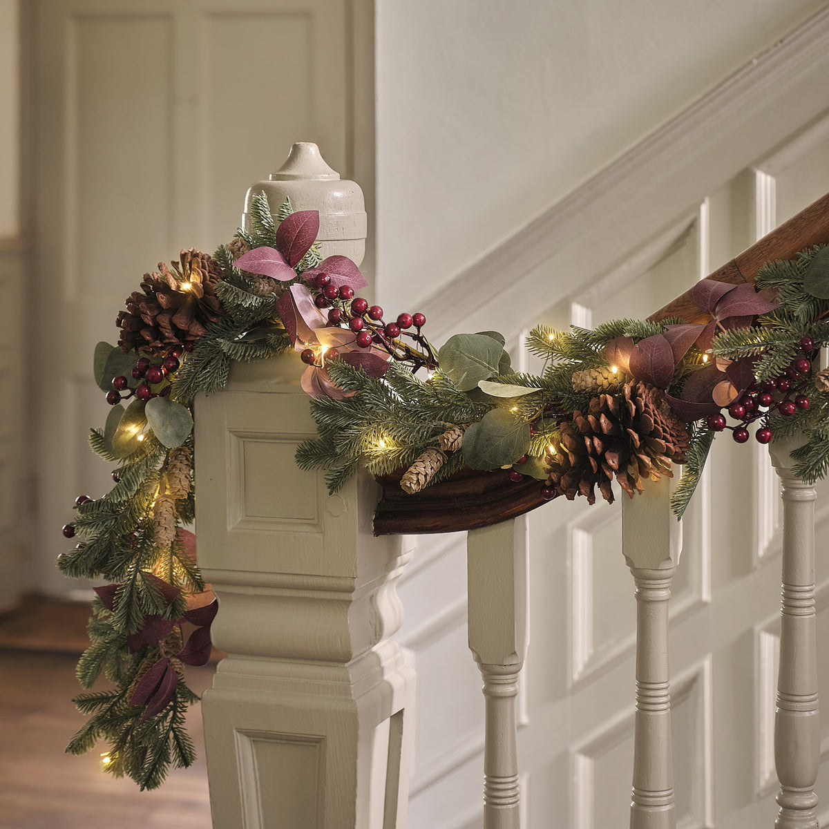 Nahaufnahme der beleuchteten Weihnachtsgirlande mit Tannenzapfen und roten Beeren am weißen Treppengeländer