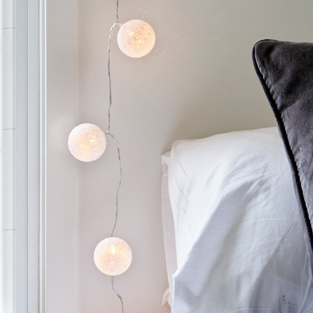 Lichterkette mit drei warmweißen LED-Baumwollkugeln neben einem Bett mit weißem Kissen