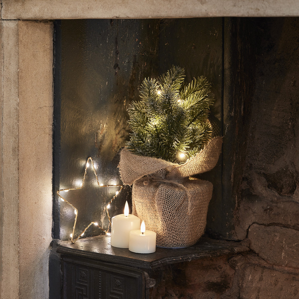 Beleuchteter Mini Weihnachtsbaum im Jutebeutel neben Kerzen und Stern-Lichterkette auf dunklem Untergrund
