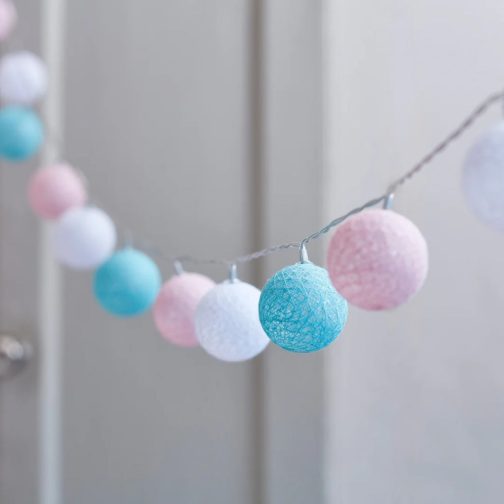 Lichterkette mit pastellfarbenen Stoffbällen in Blau, Weiß und Rosa an dünnem Kabel hängend