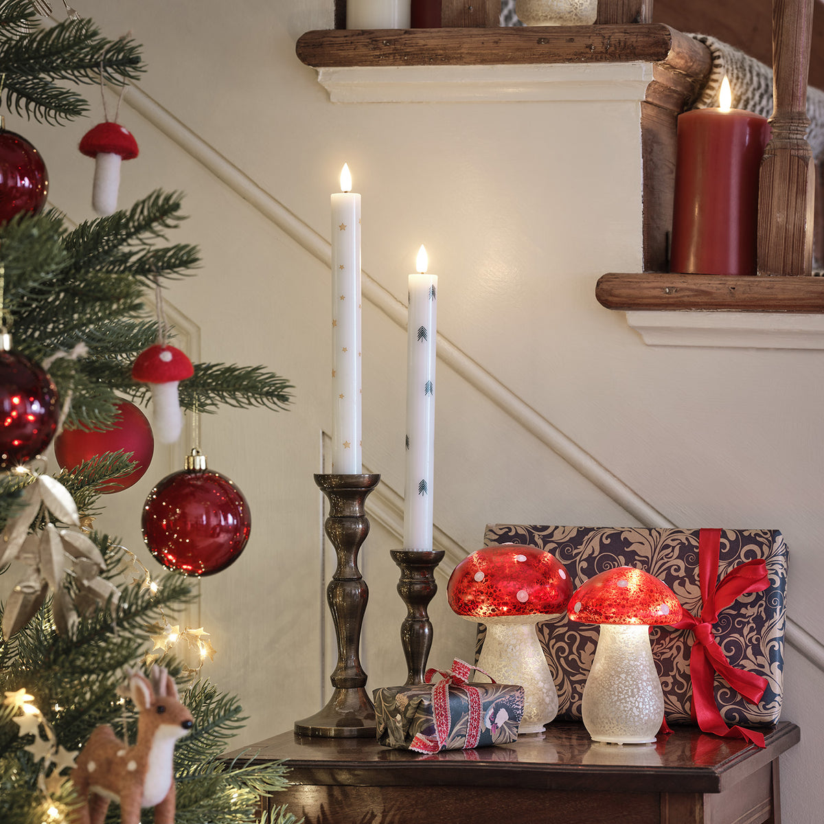 Zwei Glas-Fliegenpilze neben Kerzen und Weihnachtsgeschenken auf einem Holztisch.