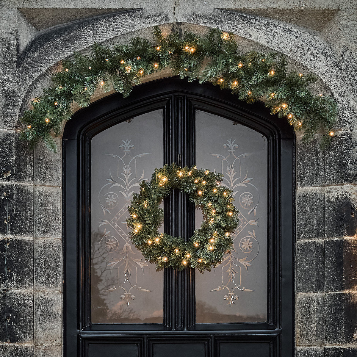 Nahaufnahme eines beleuchteten Weihnachtskranzes und einer Girlande über einer schwarzen Eingangstür