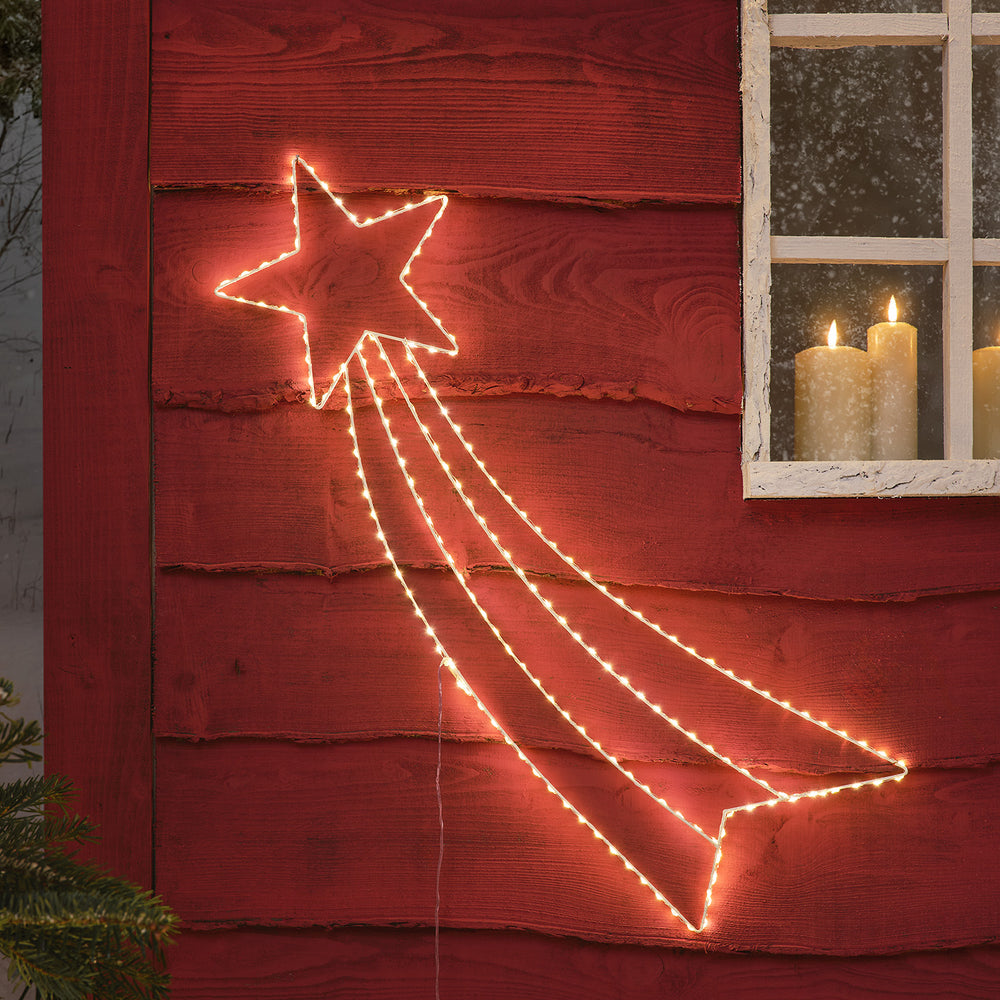 Leuchtende Micro LED Sternschnuppe an roter Holzfassade neben Fenster mit Kerzen