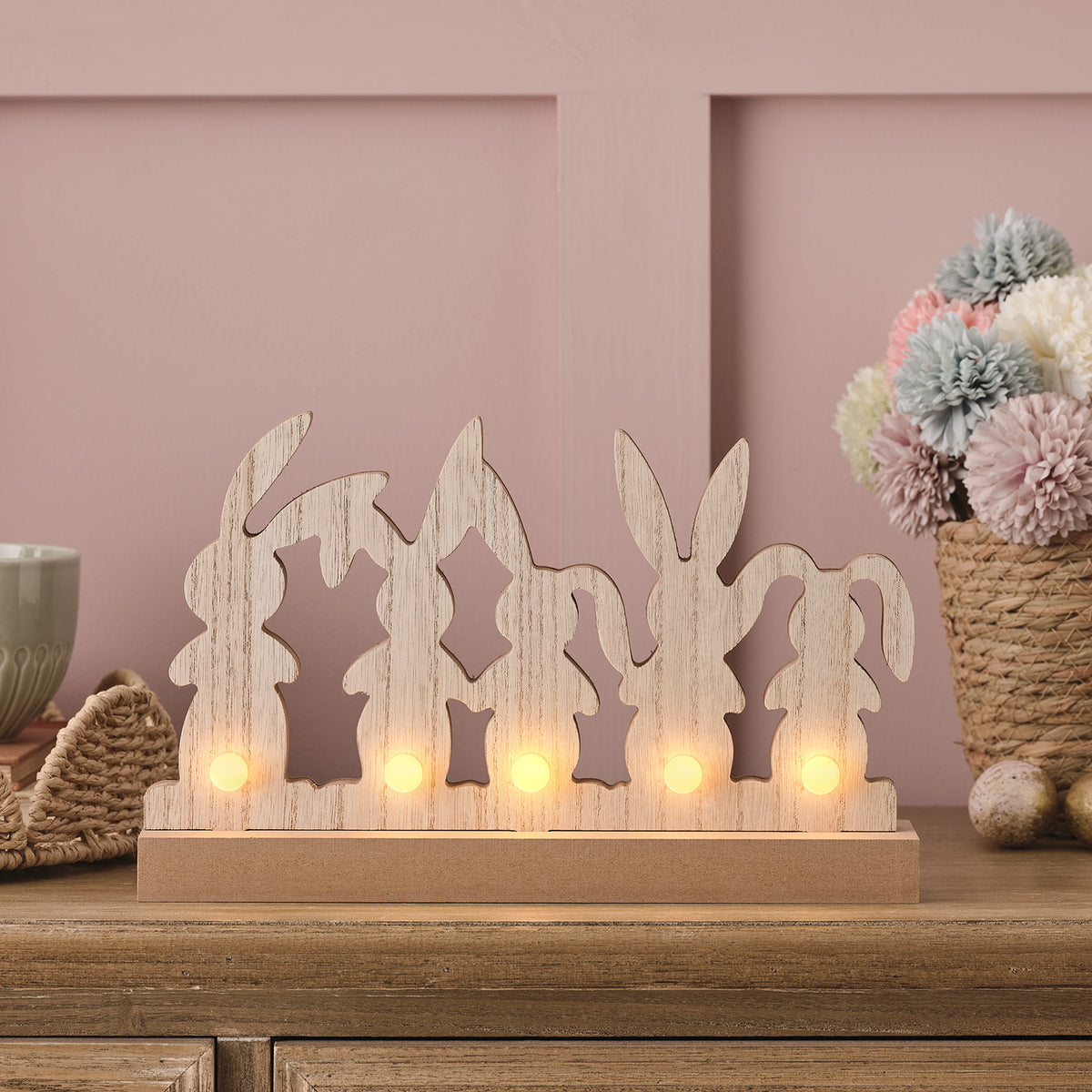 Holzdeko mit fünf Hasenfiguren und fünf warmweißen LEDs auf Holzsockel vor rosa Wand
