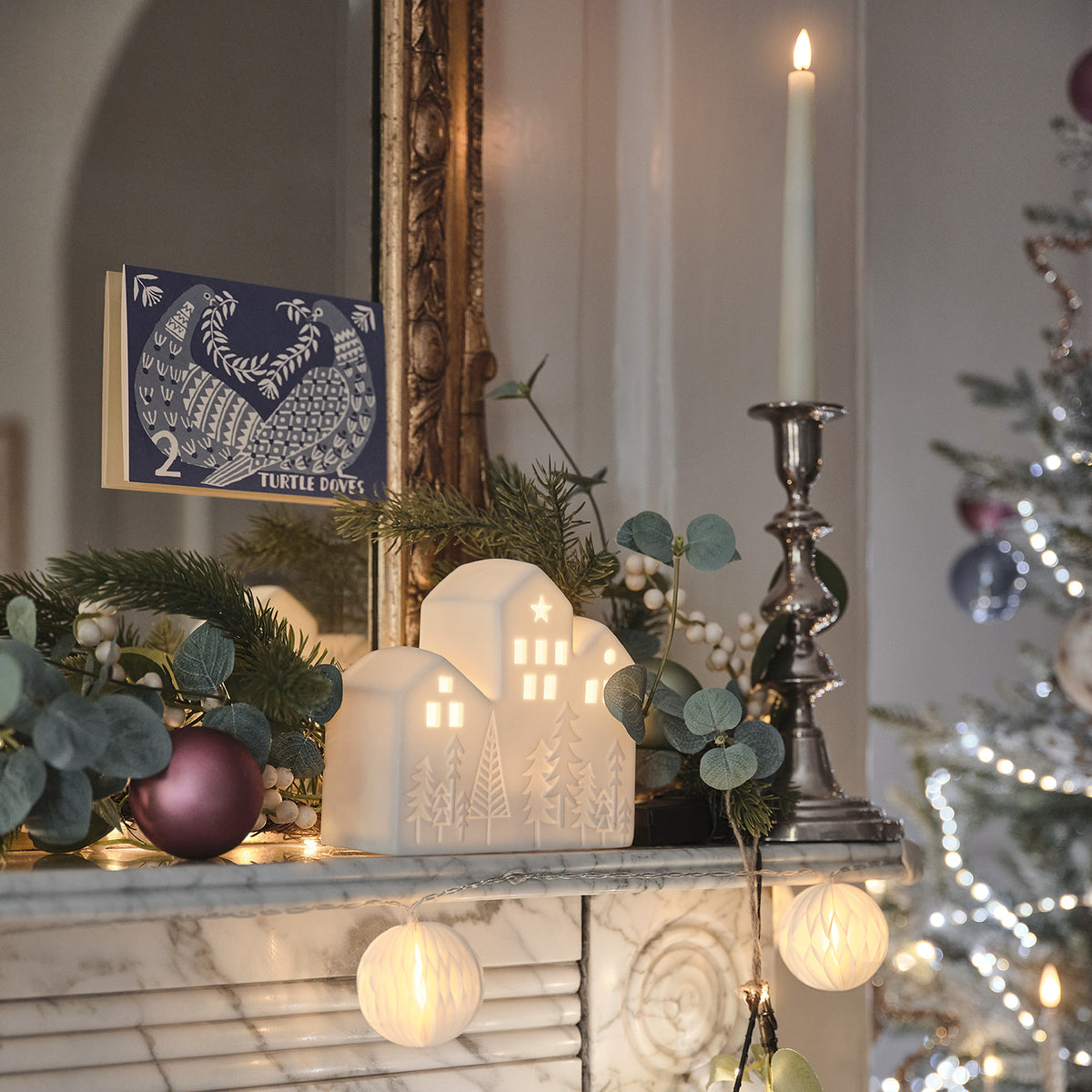 Keramik-Weihnachtshaus mit warmweißem Licht auf Kaminsims mit Weihnachtsdeko