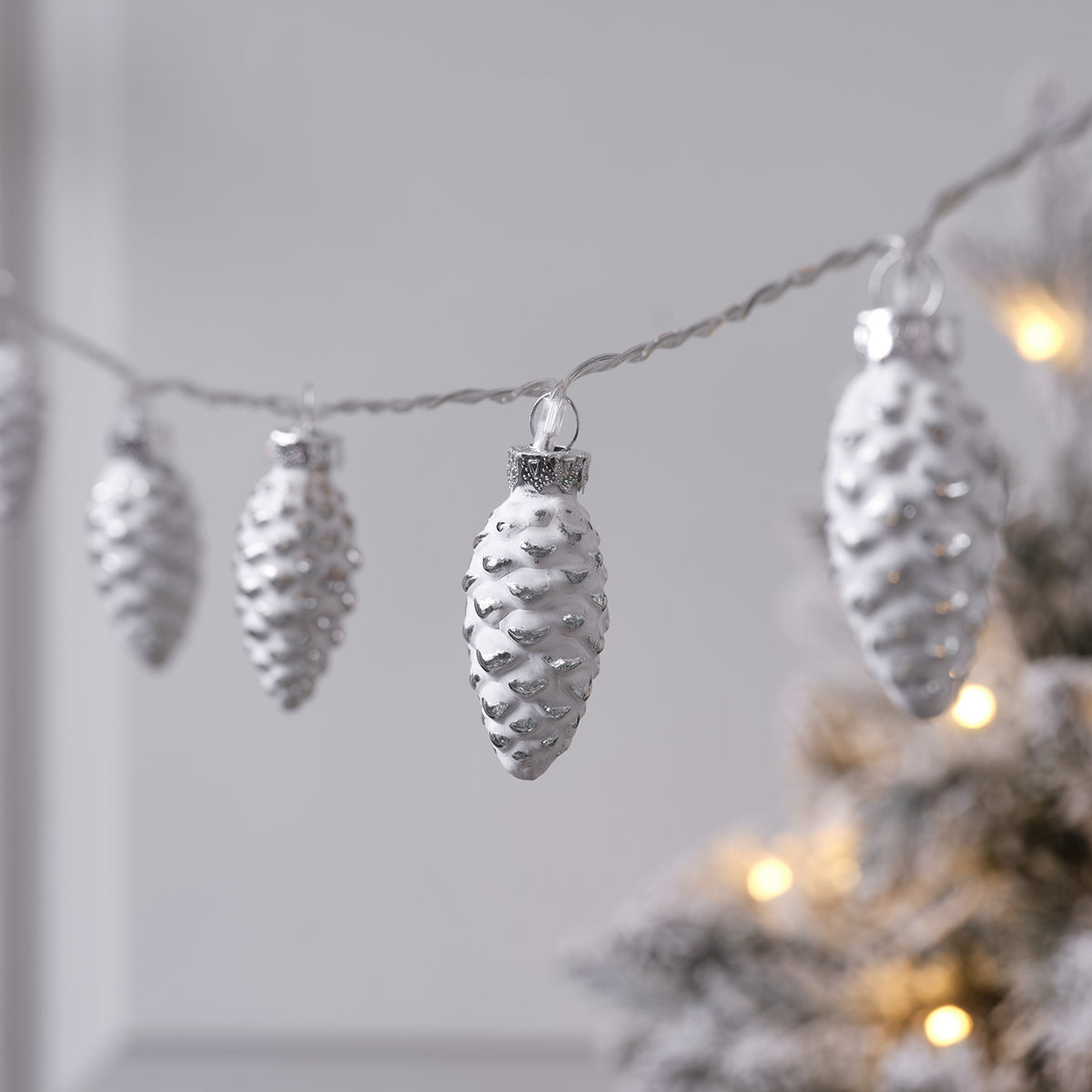 Glas-Tannenzapfen mit silbrig glänzendem Finish an Lichterkette vor hellem Hintergrund