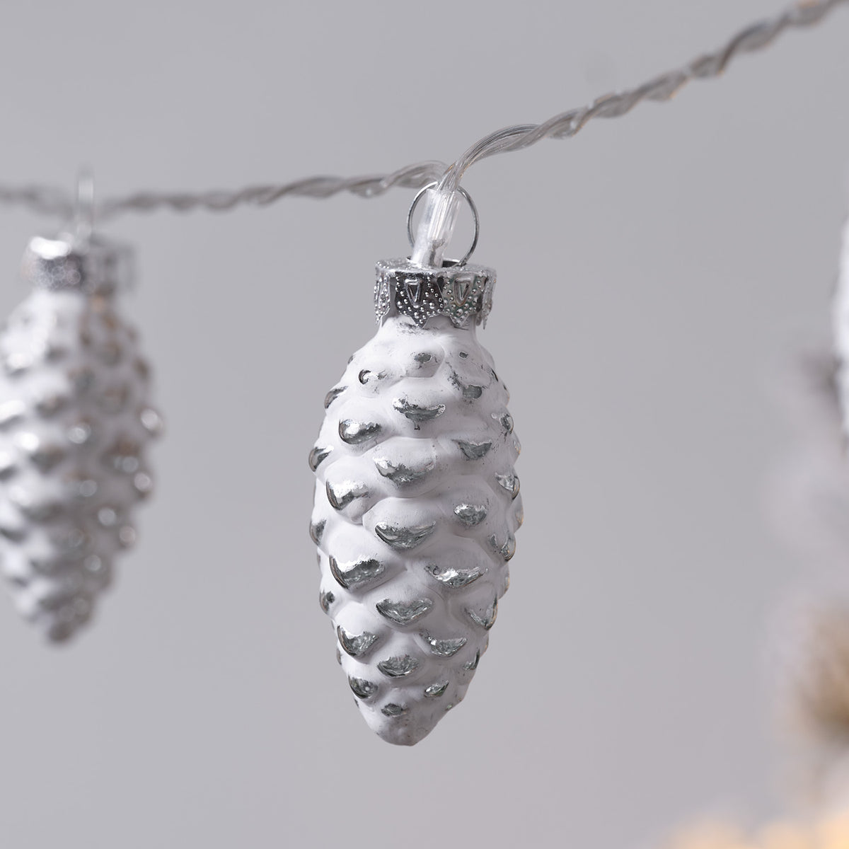 Nahaufnahme eines weißen Glas-Tannenzapfens mit silbernen Details an Lichterkette