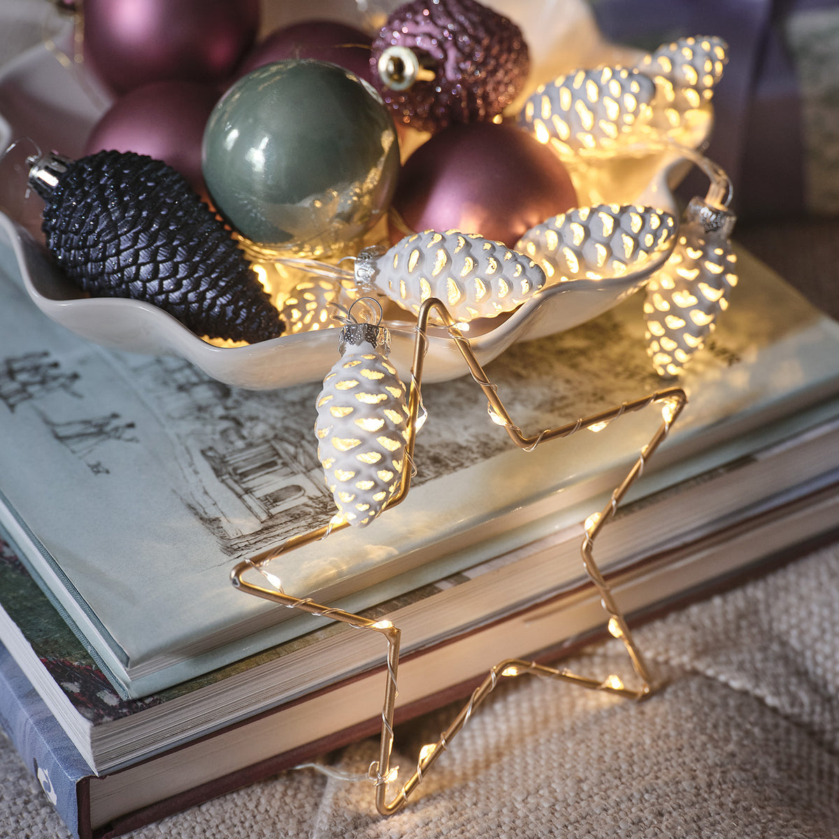 LED-Lichterkette mit weißen Glas-Tannenzapfen und Weihnachtskugeln in Schale auf Büchern