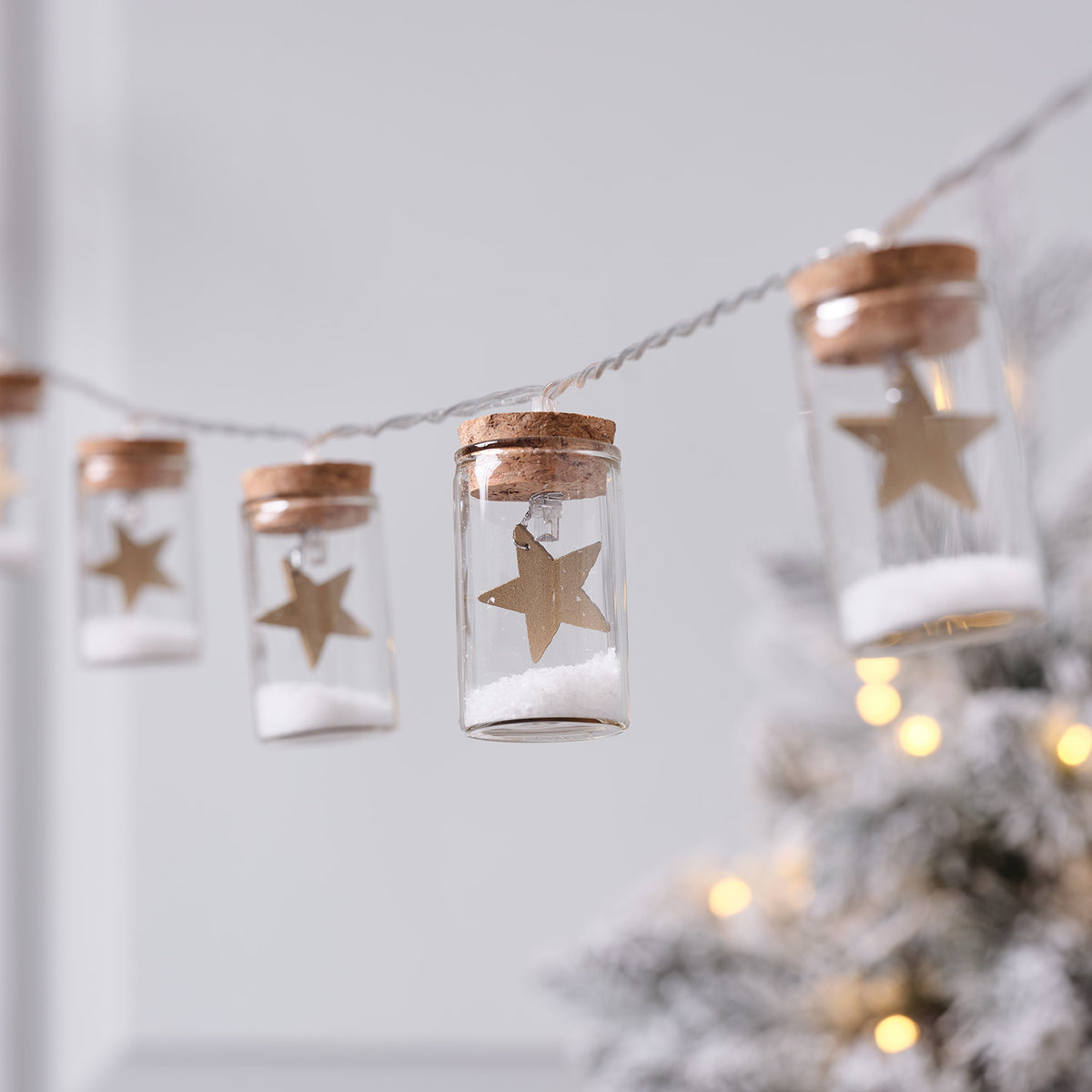 Glasfläschchen mit goldenem Stern und Kunstschnee an LED-Lichterkette vor Weihnachtsbaum