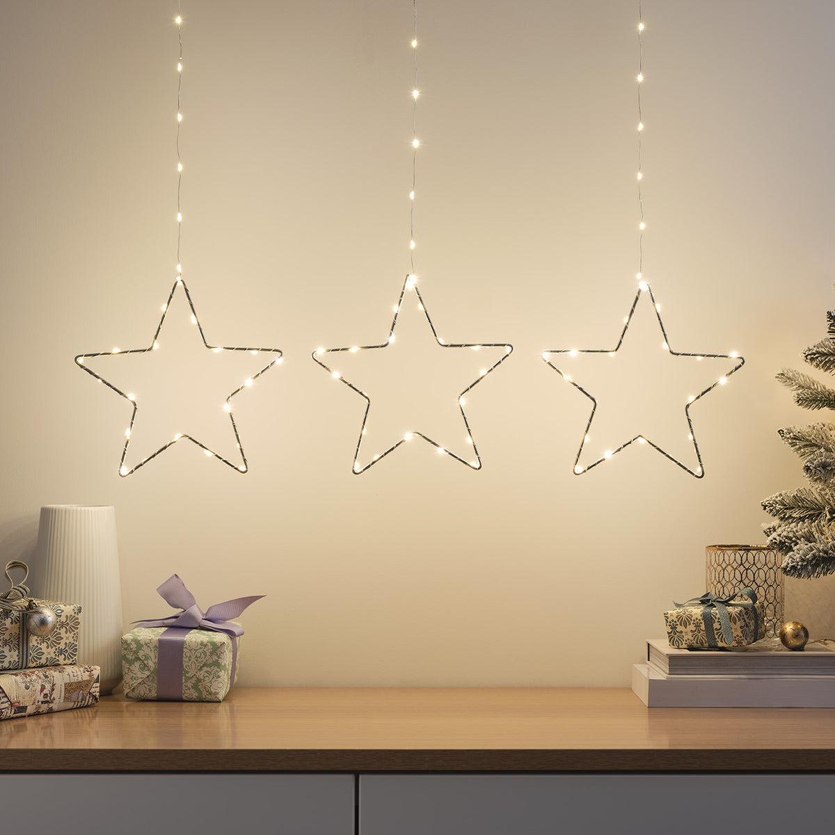 Drei leuchtende LED-Sterne aus Draht hängen vor heller Wand über weihnachtlich dekoriertem Sideboard