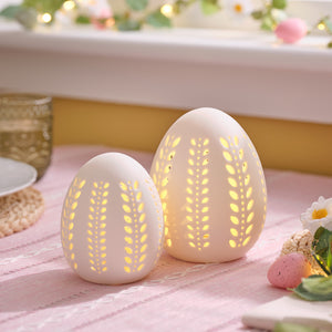 Zwei weiße Porzellan-Eier mit filigranen Blattmustern und warmweißem LED-Licht auf rosa Tischdecke