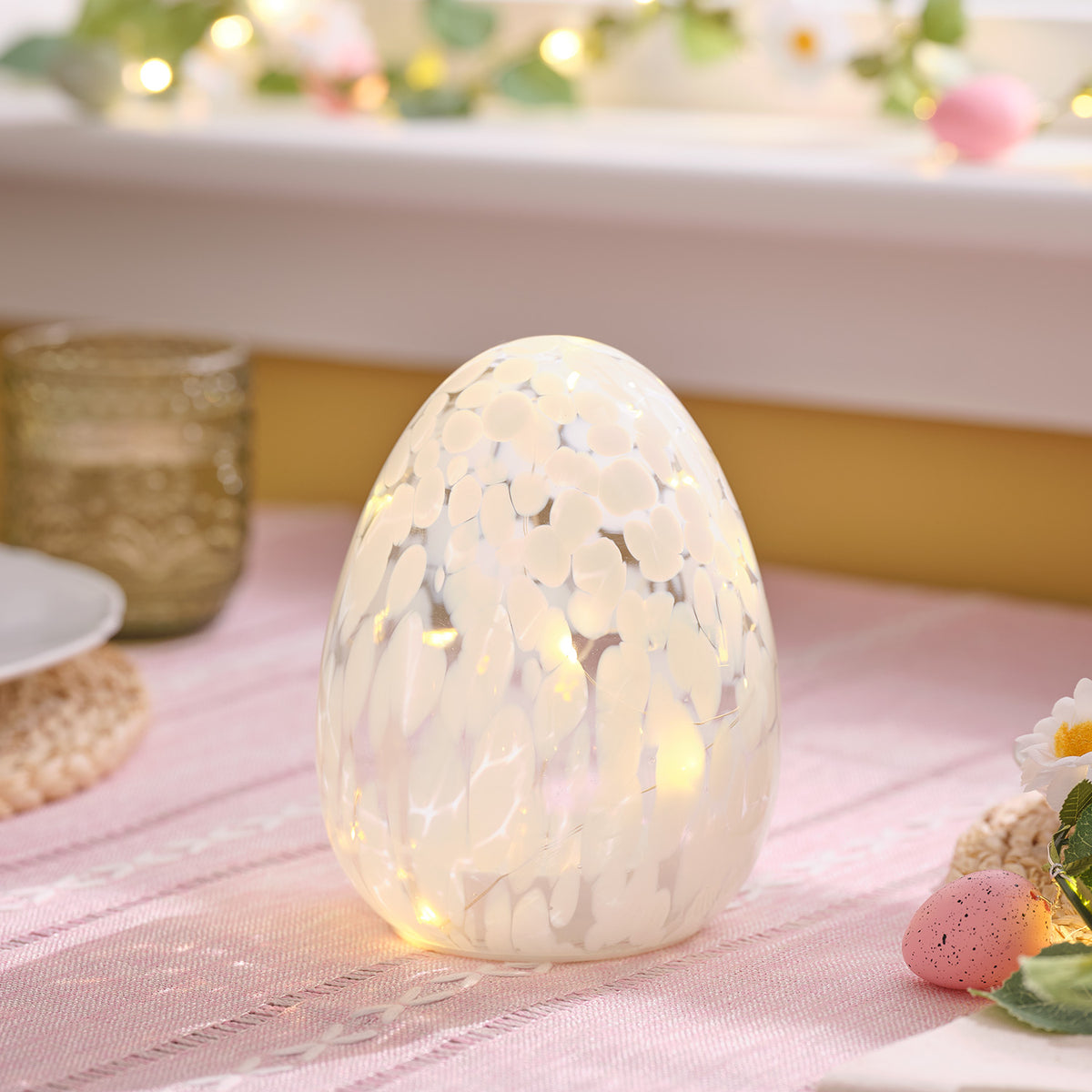 Leuchtendes LED-Osterei aus Glas mit weißem Konfettieffekt auf rosa Tischdecke