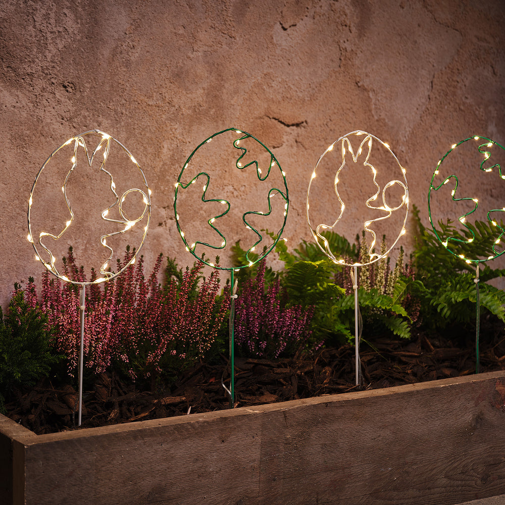 Vier LED-Osterei-Gartenstecker mit warmweißen Lichtern in einem Beet mit Pflanzen