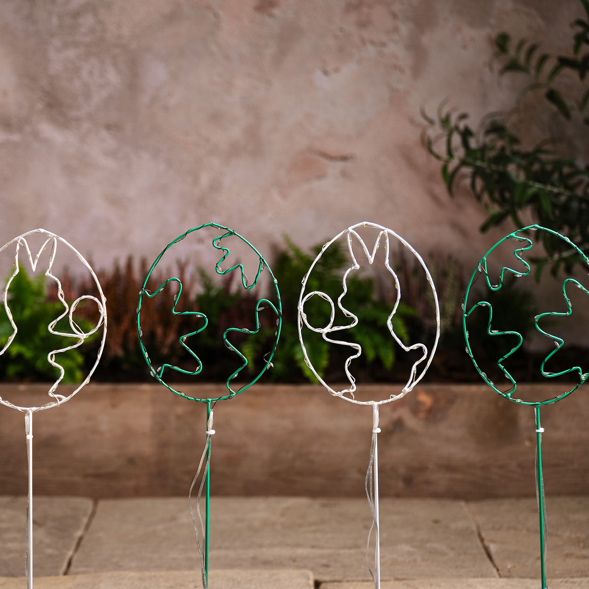 Vier unbeleuchtete LED-Osterei-Gartenstecker mit Hasen- und Blumenmotiven vor Wand