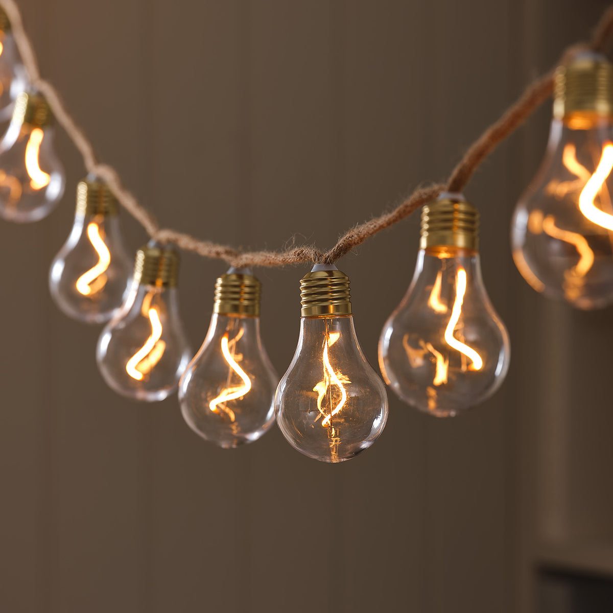 LED-Glühbirnen-Lichterkette mit warmweiß leuchtenden Birnen an Naturseil vor braunem Hintergrund