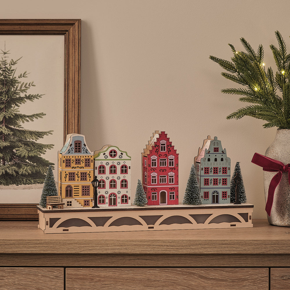 LED-Winterdorf aus Holz mit vier bunten Häuschen und schneebedeckten Bäumen ohne Beleuchtung auf Sideboard