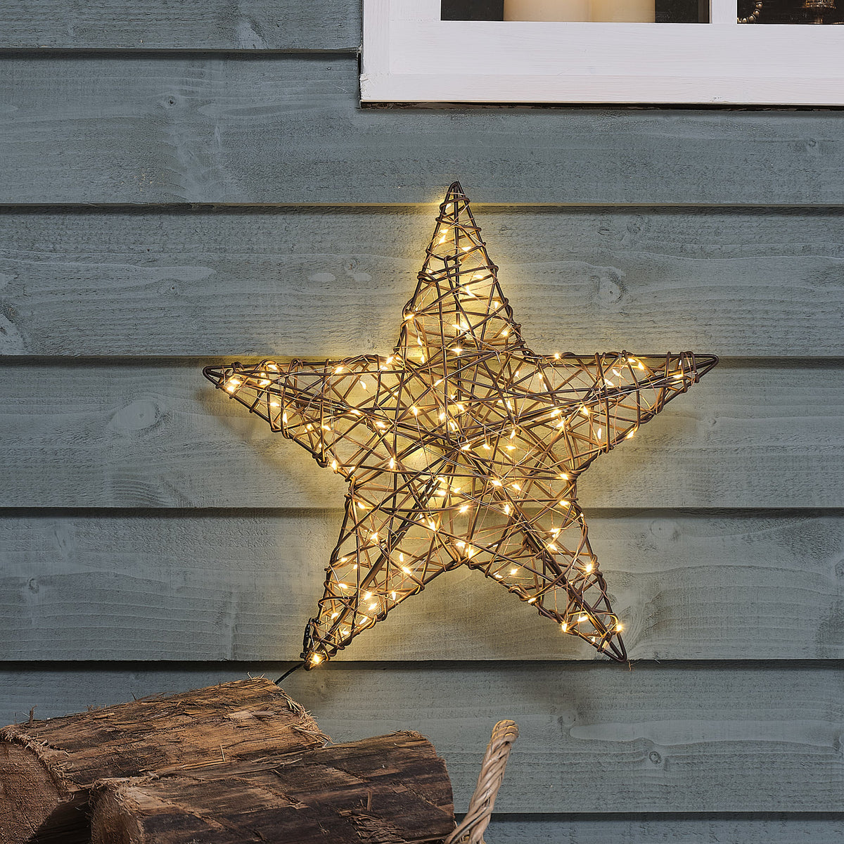 Beleuchteter großer Rattan-LED-Stern 40 cm an grauer Holzfassade mit Holzscheiten im Vordergrund