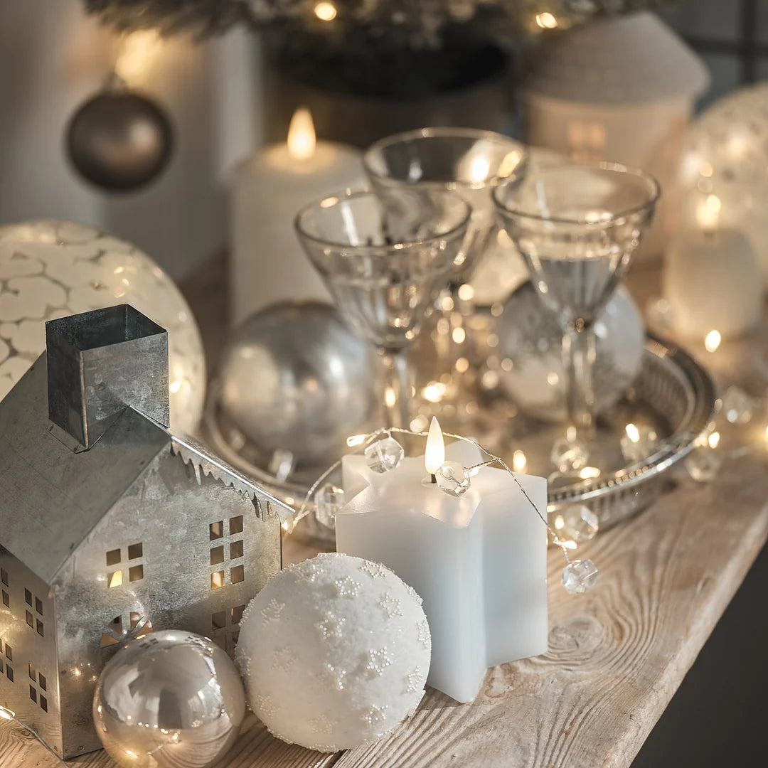 Festliches Weihnachtsarrangement mit Metall-LED-Weihnachtshaus, weißer sternenförmiger LED-Kerze, Schneekugeln und Sektgläsern im Hintergrund.