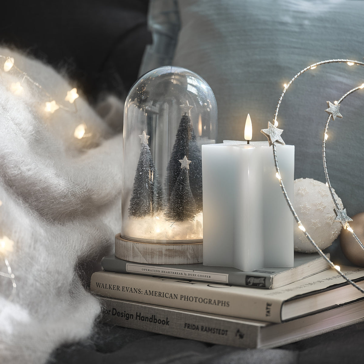 LED-Glasglocke mit Weihnachtsbäumen neben weißen Sternkerzen und Weihnachtskugel auf Büchern