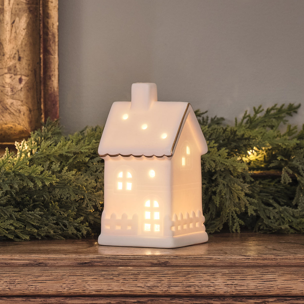 Weißes LED-Keramikhaus mit warmweißem Licht auf Holzfläche vor grünen Zweigen