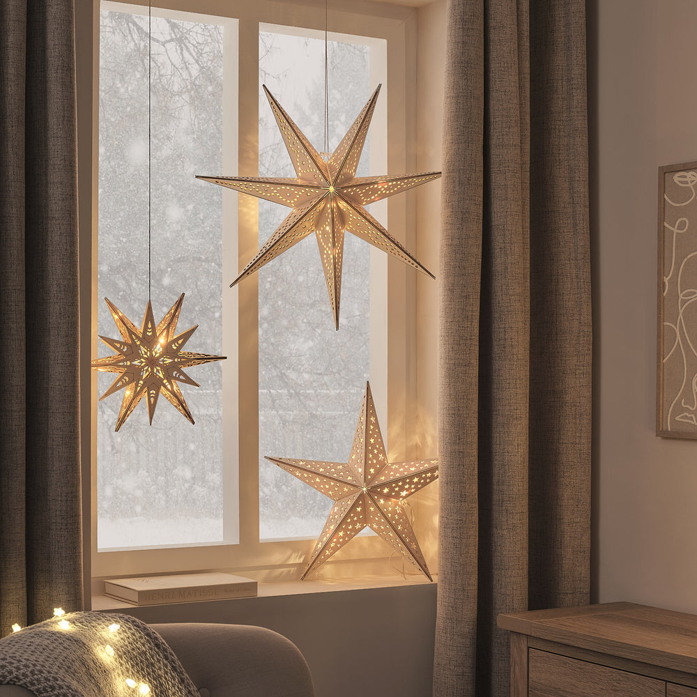 Beleuchtete goldene Holzsterne mit verschiedenen Zacken hängen vor einem Fenster mit Vorhängen