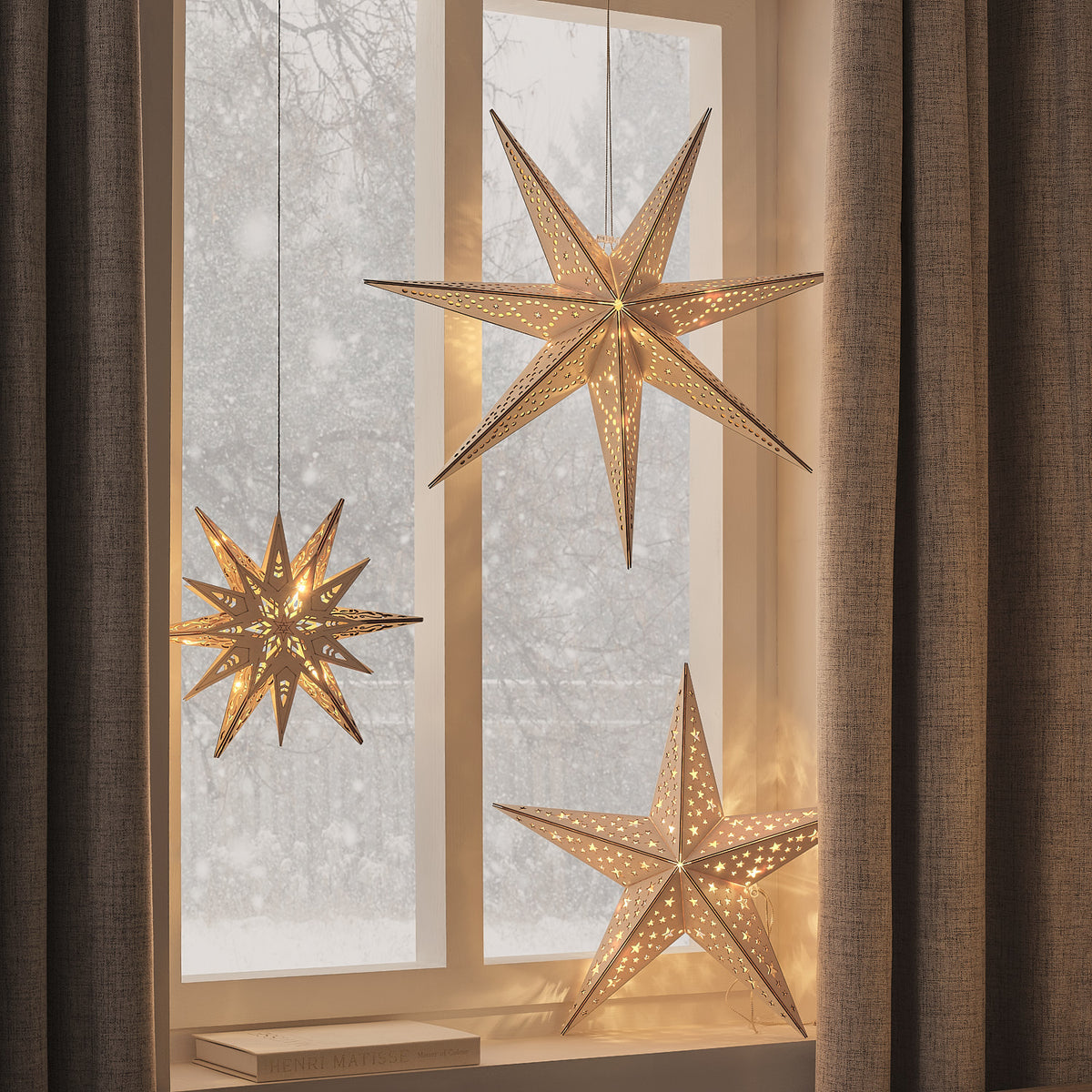 Goldene LED-Holzsterne mit warmweißem Licht hängen vor einem Fenster mit braunen Vorhängen