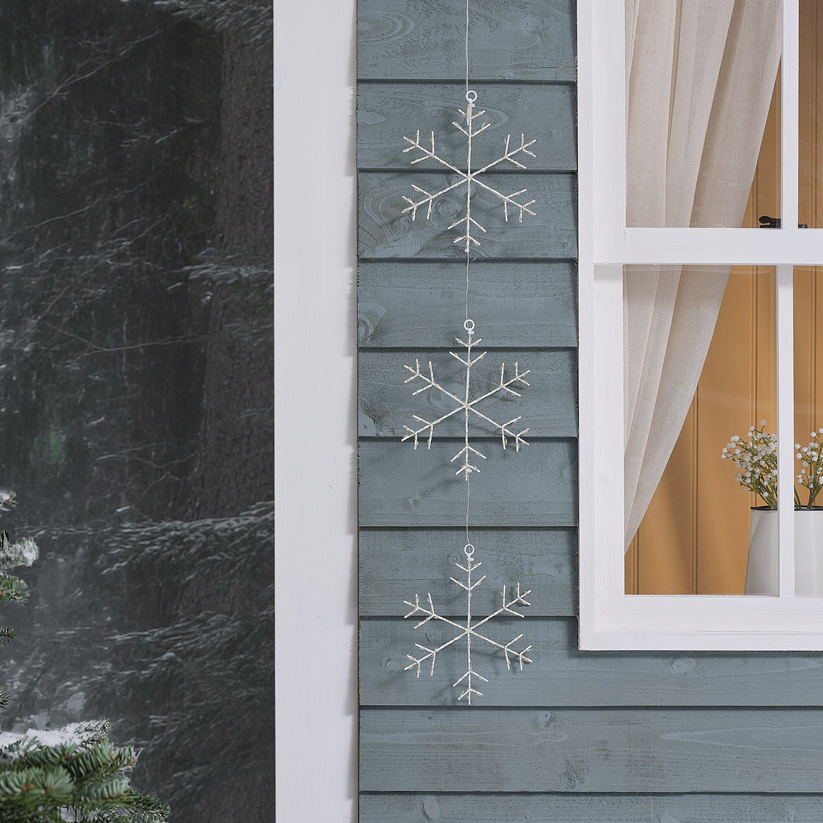 Drei unbeleuchtete LED-Schneeflocken an einer grauen Holzfassade neben einem Fenster