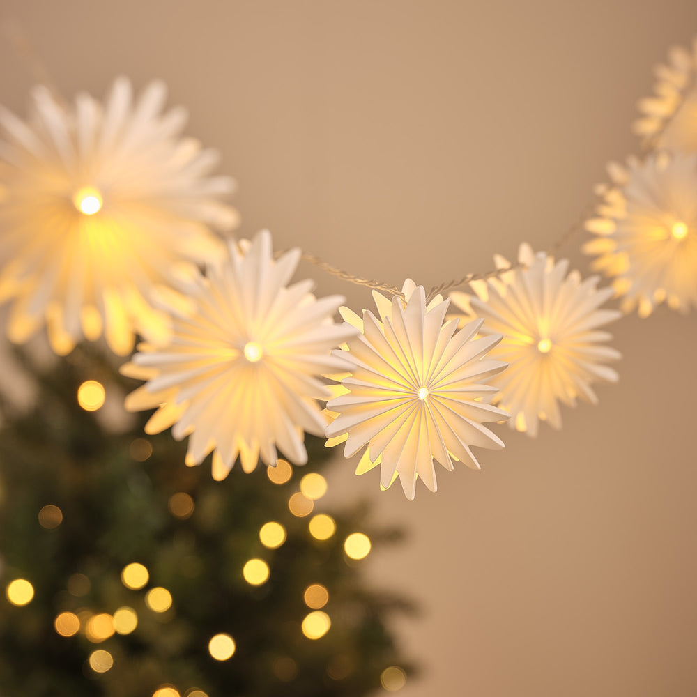 LED-Lichterkette mit zehn warmweiß leuchtenden Papier-Schneeflocken vor unscharfem Hintergrund