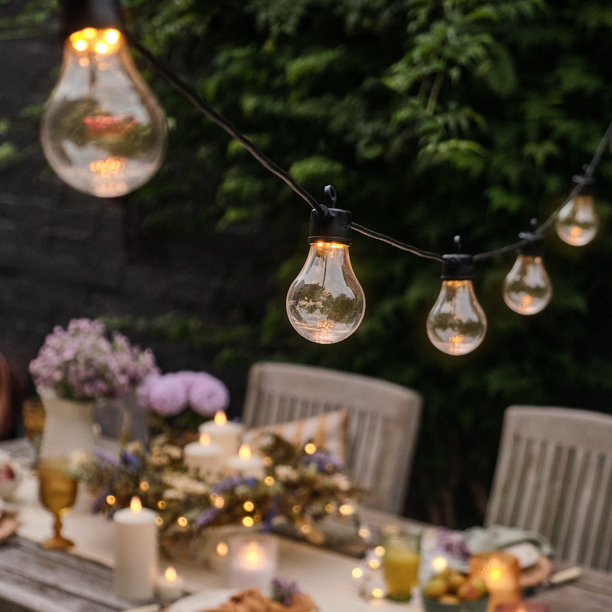 Nahaufnahme einer warmweißen Glühbirne an schwarzer Lichterkette über gedecktem Gartentisch