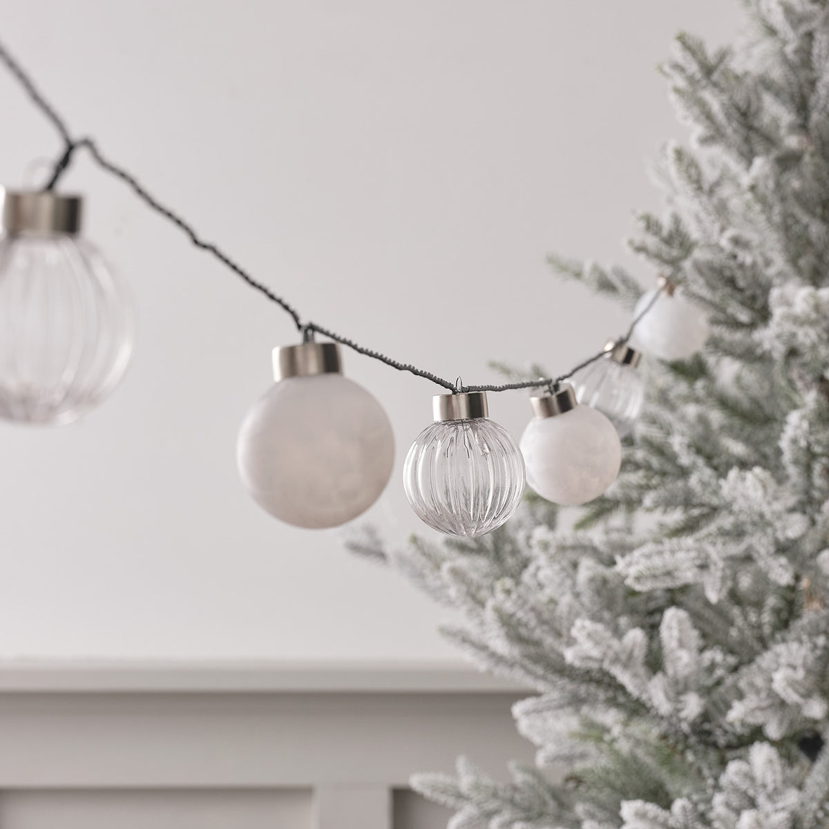 LED Lichterkette mit weißen und klaren Weihnachtsbaumkugeln an grünem Kabel vor Tannenzweigen