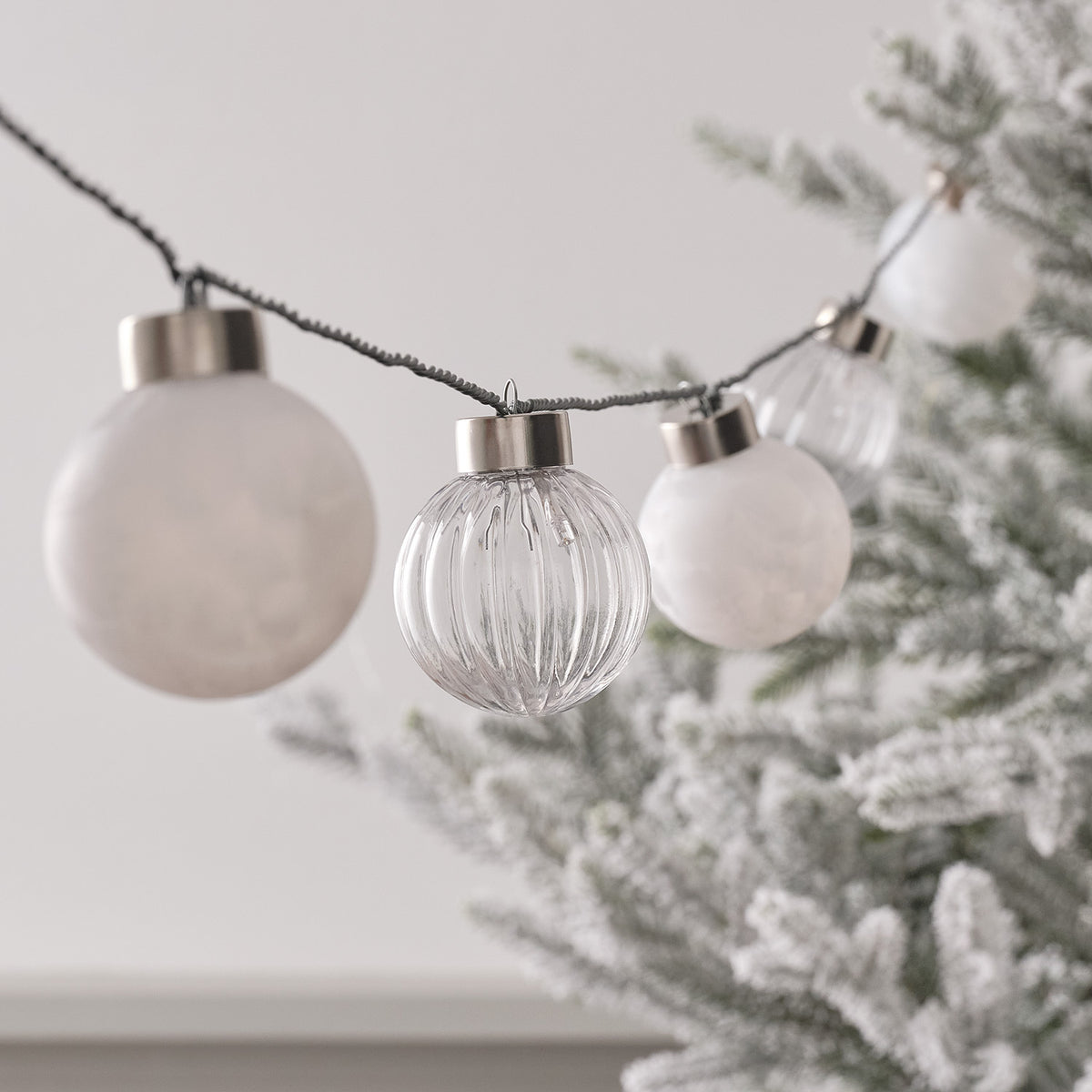 LED Lichterkette mit weißen und klaren Weihnachtsbaumkugeln an grünem Kabel vor Tannenzweigen