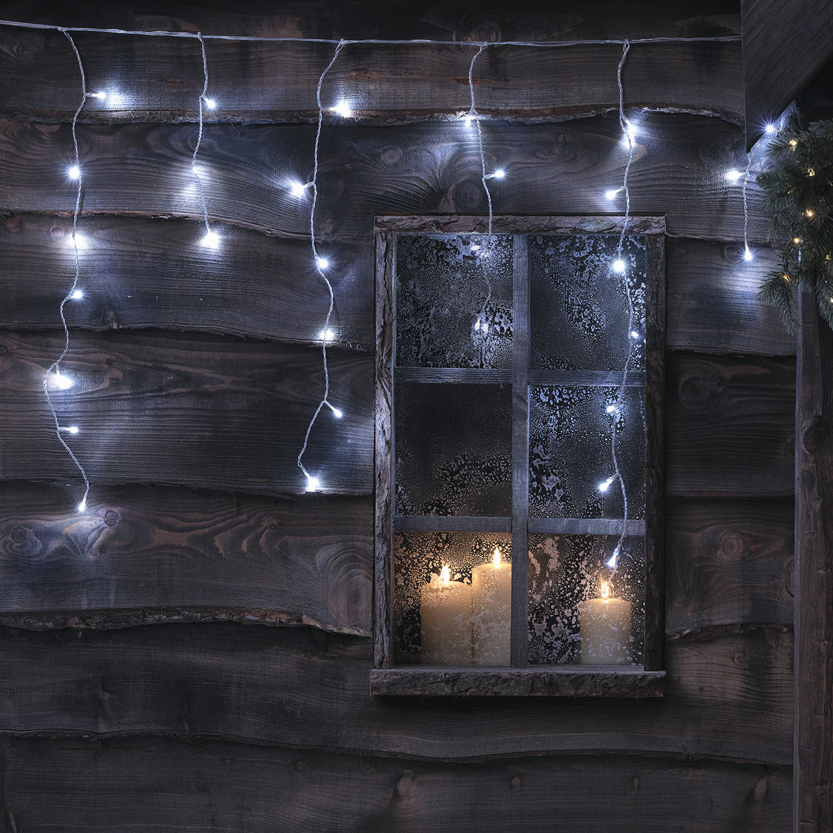 Bicolor LED Eiszapfen Lichterkette in kaltweiß an Holzfassade mit Fenster und Kerzen im Inneren