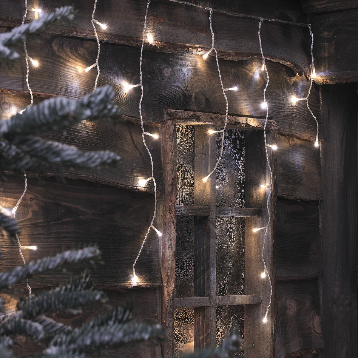 Bicolor LED Eiszapfen Lichterkette in warmweiß an Holzfassade mit Fenster und Kerzen im Inneren