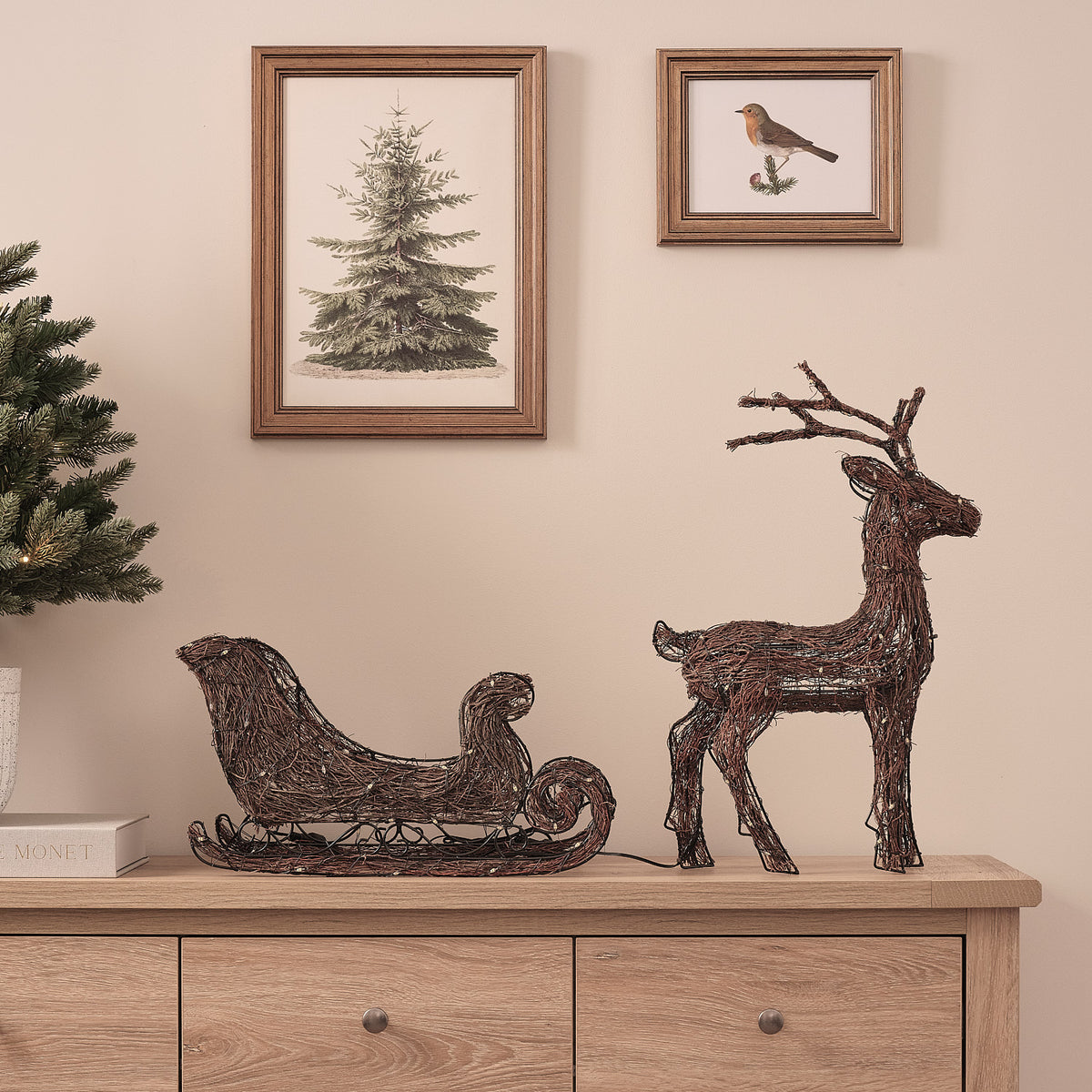 LED-Rentier mit Schlitten aus Naturzweigen ohne Beleuchtung auf Kommode vor Wand mit Bildern